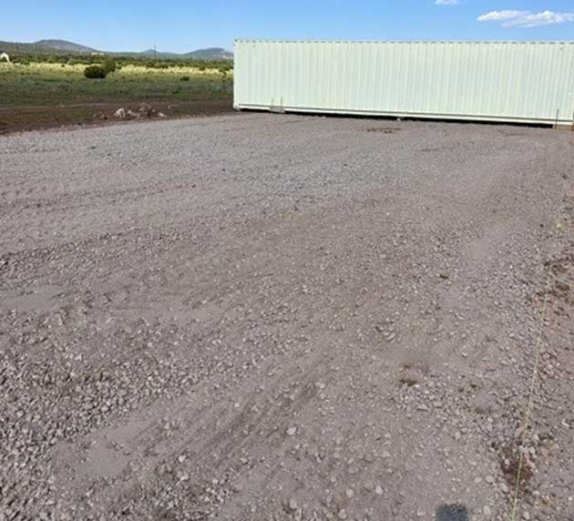 A long, light-colored shipping container sits on a gravel lot, with a grassy field and hills in the background under a blue sky.