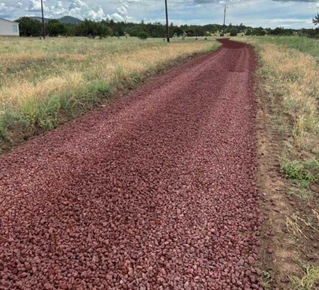 A gravel road made of reddish stones stretches through a grassy landscape with a few distant clouds in the sky.