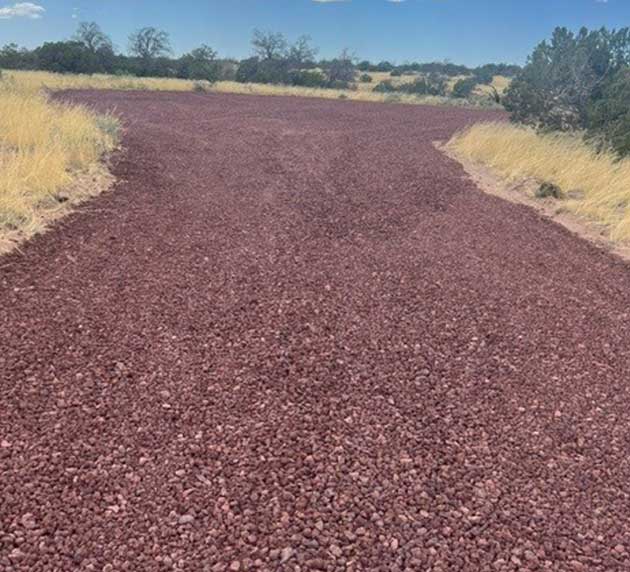A gravel path made of reddish-brown stones stretches forward, flanked by dry grass and shrubs under a blue sky.
