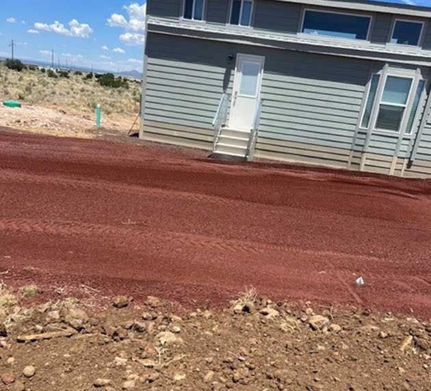 Small house with gray siding on a red dirt lot, under a clear blue sky. Sparse vegetation and utility poles are in the background.