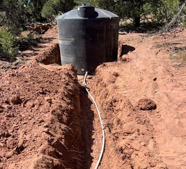 A black water tank with a pipe is partially buried in a trench in a dirt area surrounded by trees.