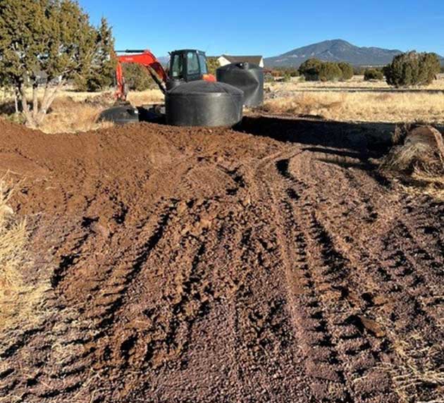 An excavator is parked on a dirt path with a large black tank beside it. The area is surrounded by dry grass and trees, with a mountain visible in the background.