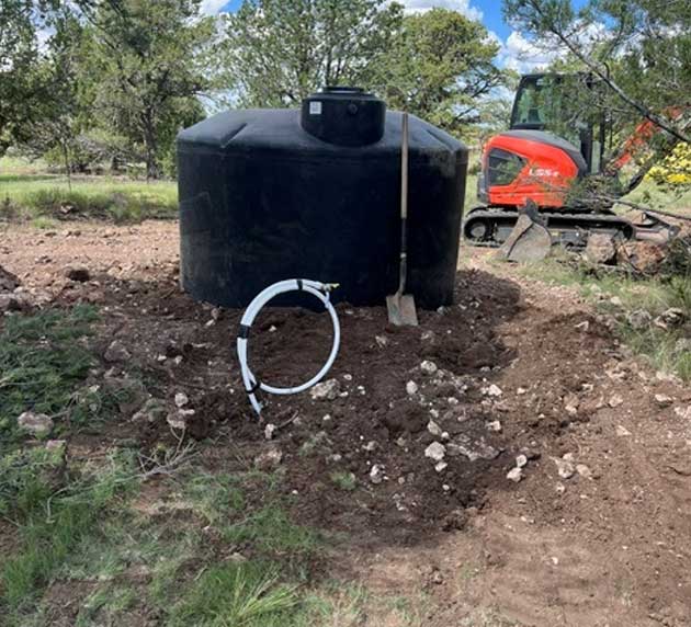 A black water tank near a pile of soil with a shovel stuck in it. A hose lies coiled nearby, and an orange excavator is visible in the background among trees.