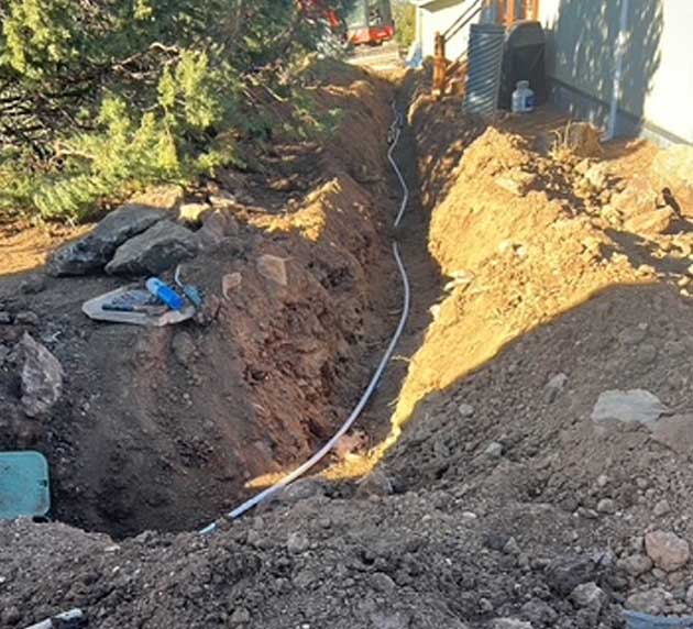 A narrow trench with exposed brown earth and a pipe running through it alongside a building.